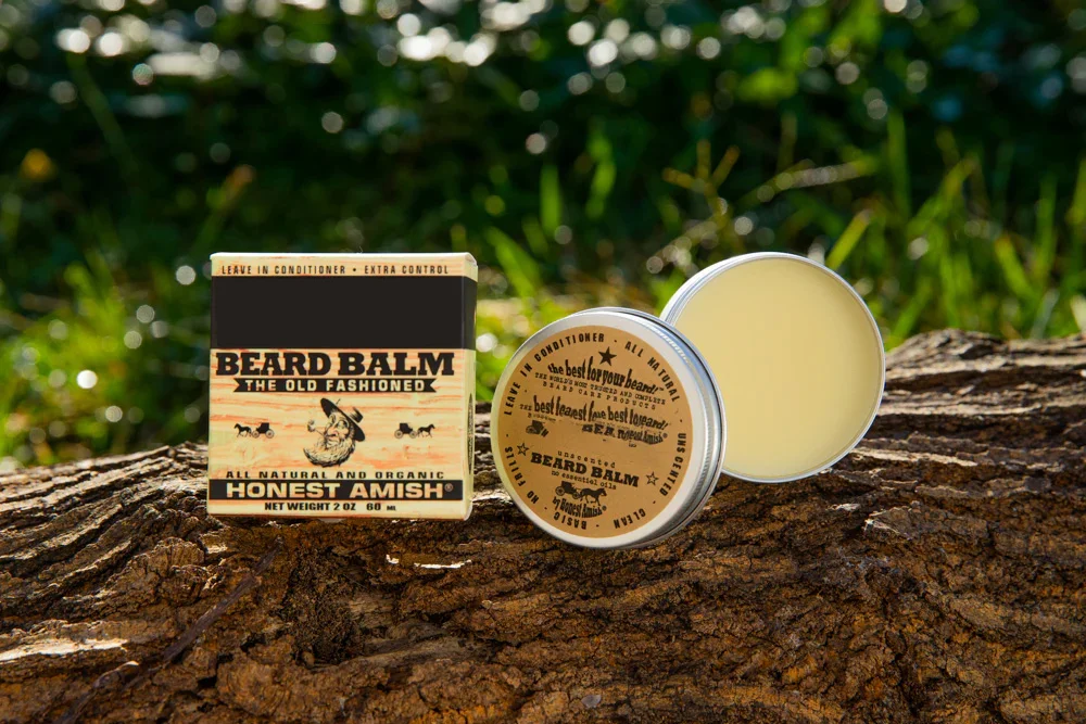 Honest Amish beard balm next to its box on a tree stump in a natural outdoor setting with greenery in the background.